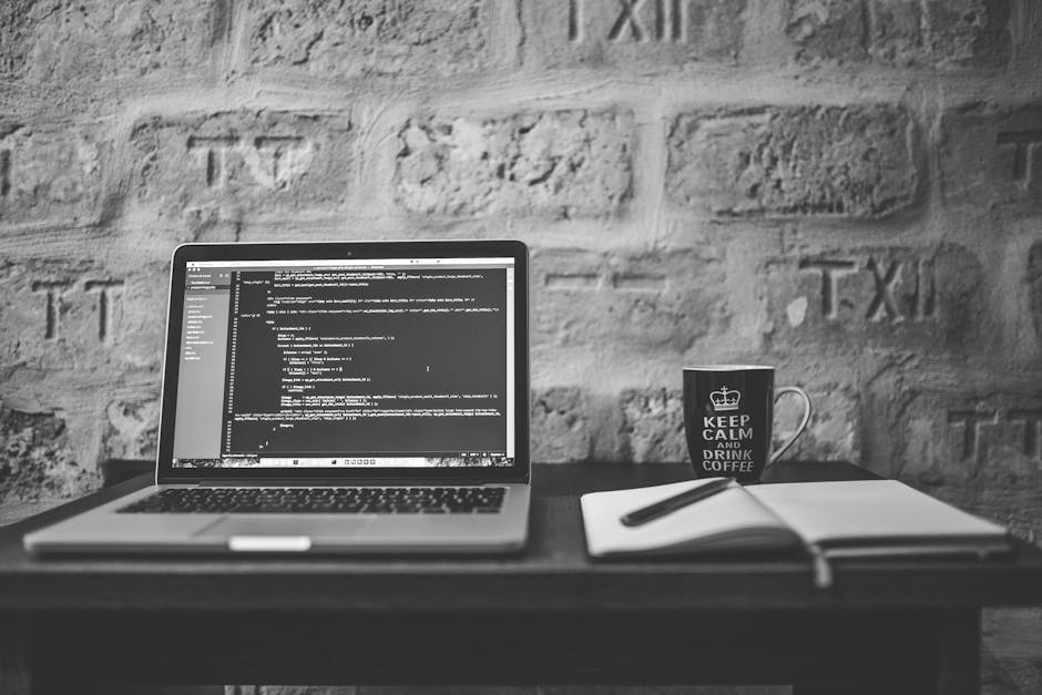 A black-and-white photograph of a workspace featuring an open laptop on a dark wooden desk displaying lines of code on the screen, with a white notebook and a pen resting on top of it. To the right of